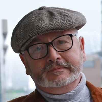 With flat cap and brown corduroy jacket, modern City offices behind.
