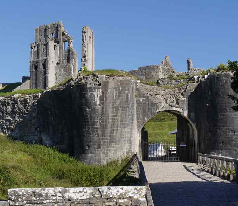 Corfe Castle Entrance gate with the ruins on hill in distance beneath a blue sky.